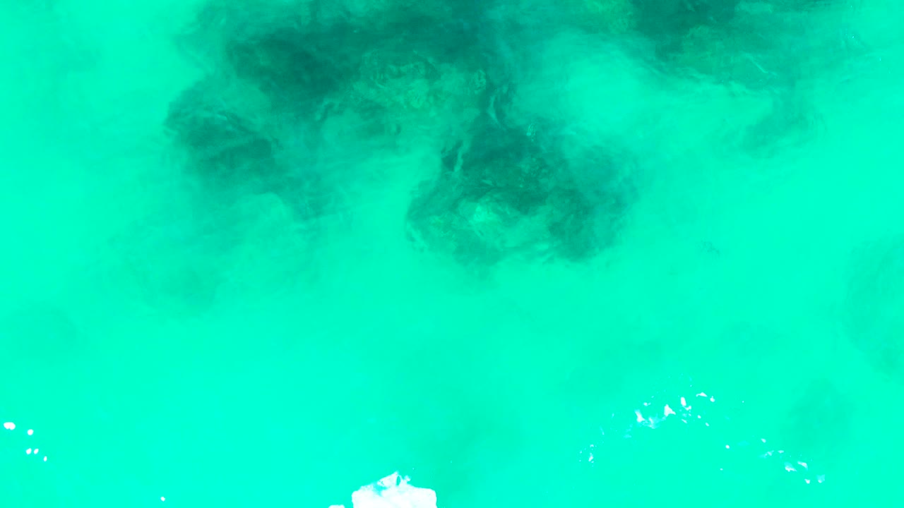 Top-down frame over Punta Buzzards water with darker submerged patches across the upper half, clear turquoise shallows, white foam entering near the lower left, and no visible people.