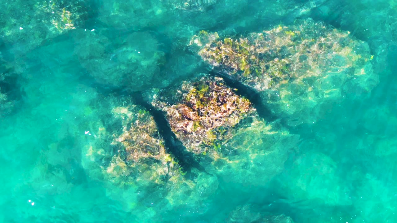 Top-down view of clear turquoise water over several submerged rocks at Punta Buzzards, Baja California Sur, with bright ripples and no visible people.