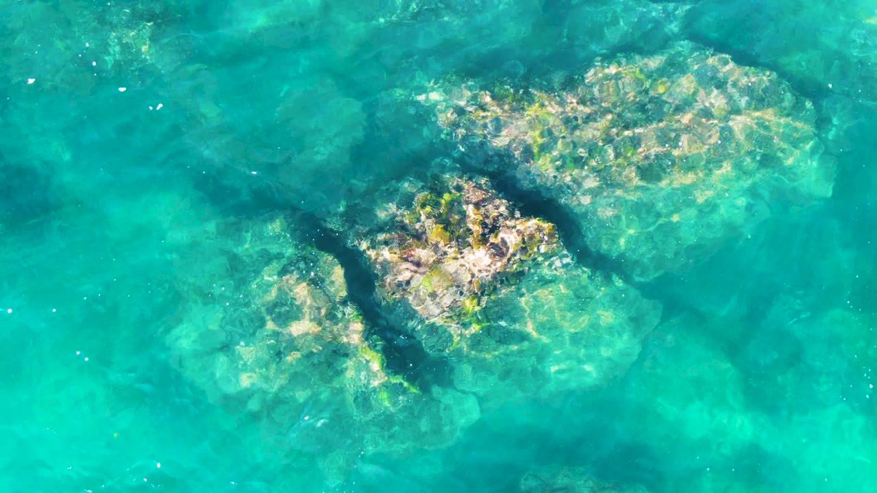 A later top-down frame shows the same shallow turquoise water at Punta Buzzards, Baja California Sur, with rock shapes slightly shifted under ripples and no visible people.
