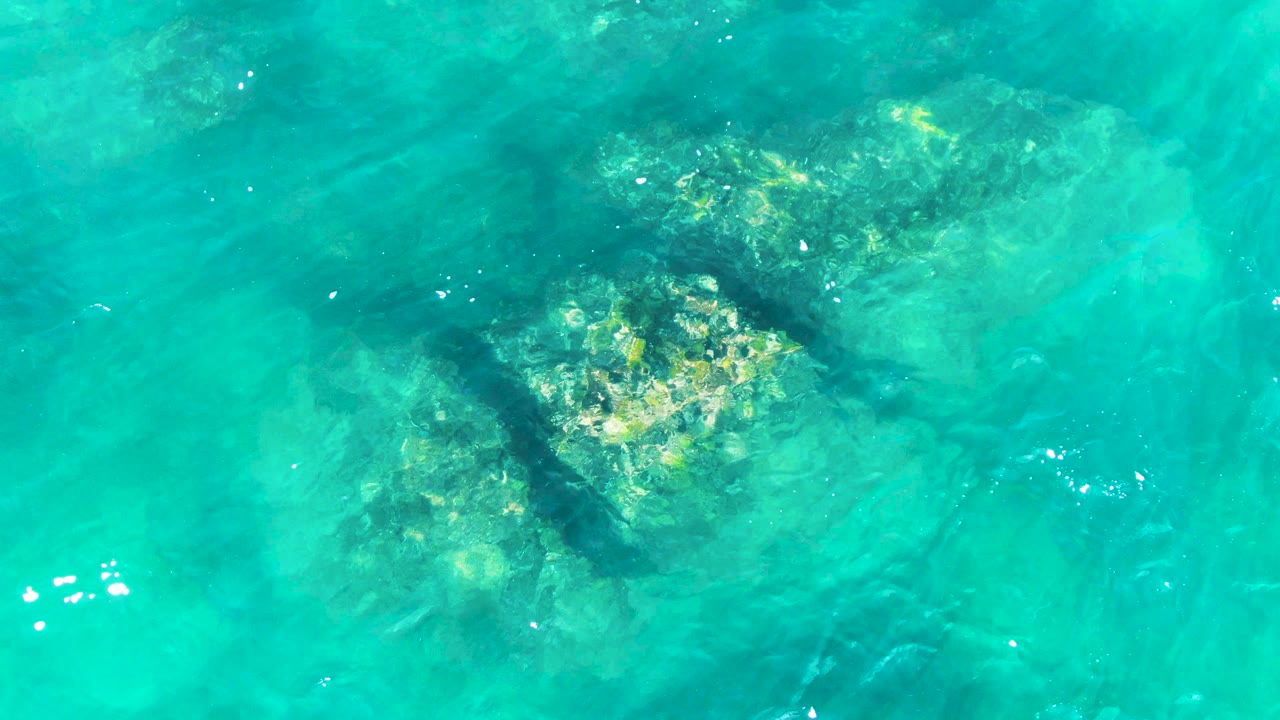 Turquoise ocean water at Punta Buzzards reveals reef-like rocks below the surface, with bright flecks of reflected light and no visible people.