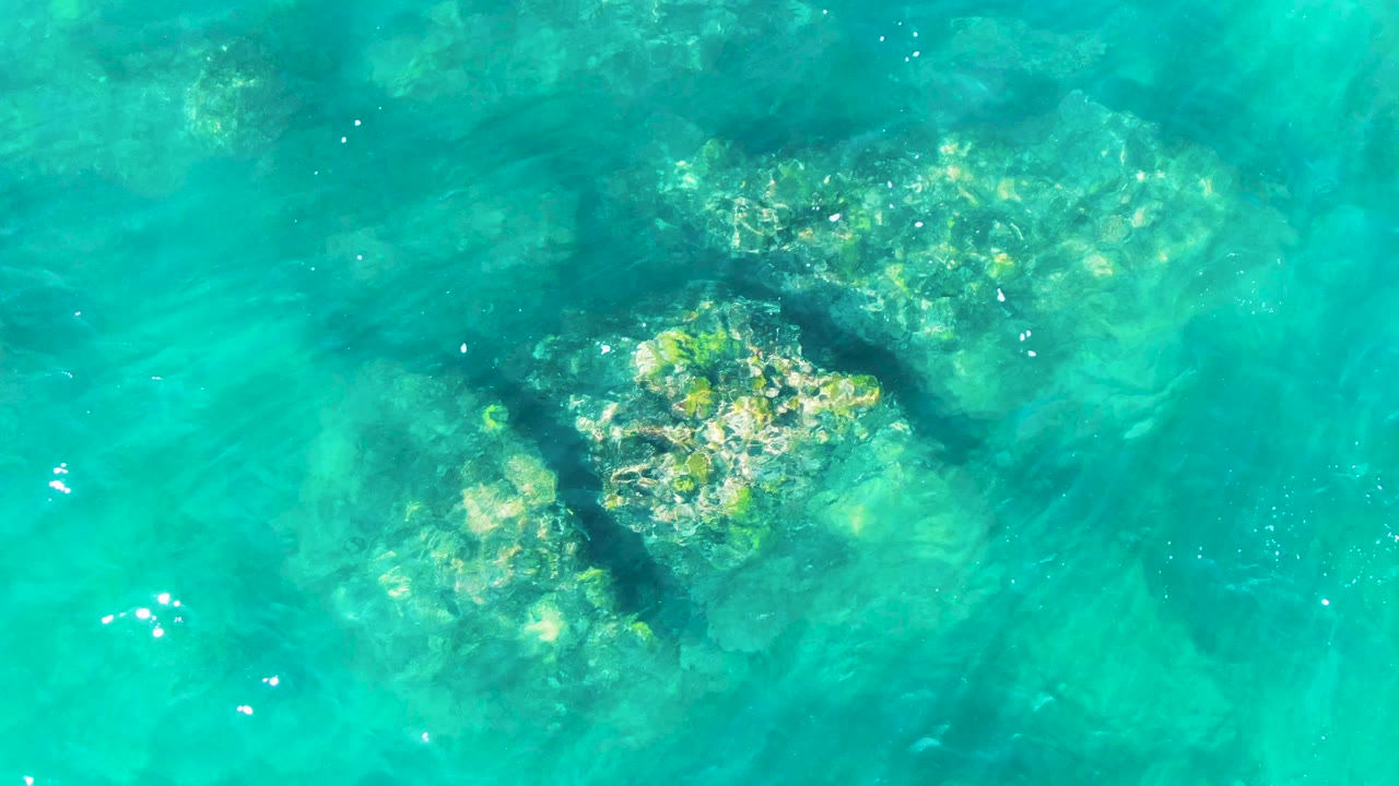 A submerged rock patch sits in clear turquoise water at Punta Buzzards, with darker water shadows around it and no visible people.
