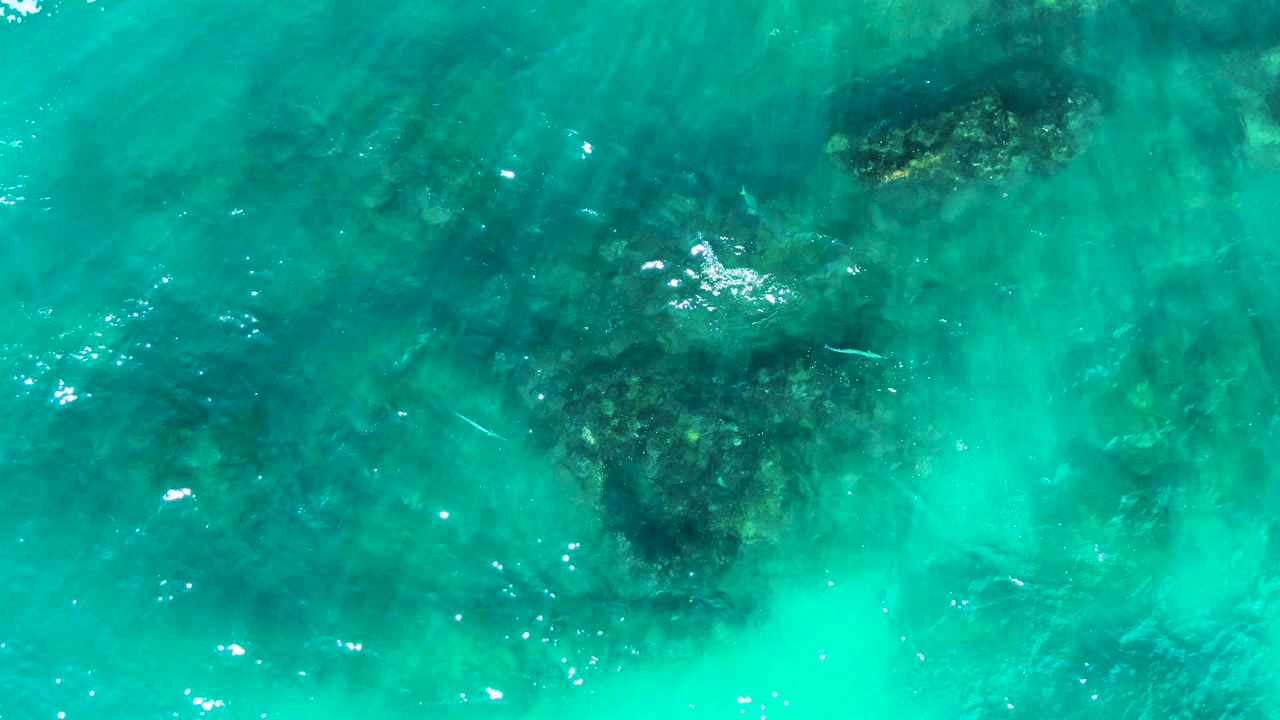 Aerial view at Punta Buzzards, Baja California Sur, with turquoise water covering a cluster of submerged rocks near the left side, scattered white reflections, and no visible people.