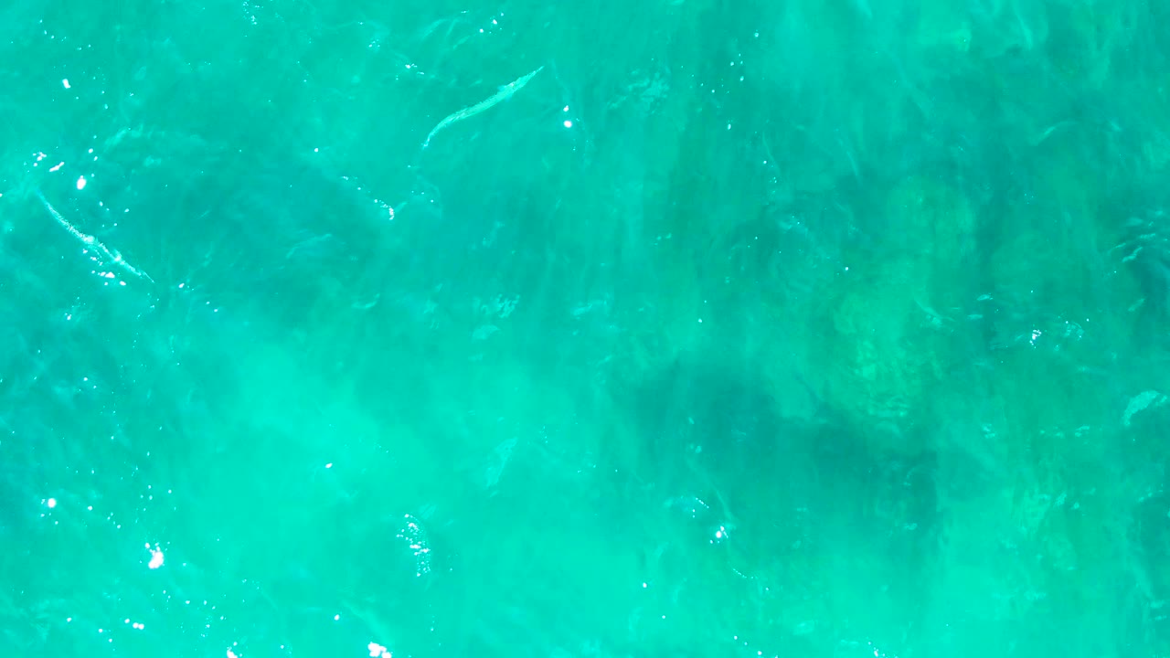 Overhead view of clear turquoise water at Punta Buzzards, Baja California Sur, with ripples, sunlight flecks, and faint dark underwater patches, no visible people.