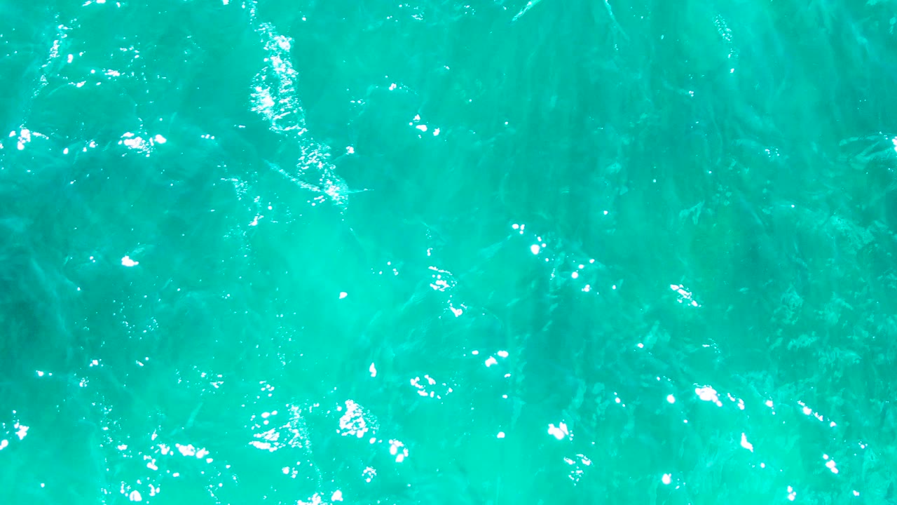 Clear blue green ocean water fills the frame at Punta Buzzards, with bright surface reflections and soft darker shapes visible below, no visible people.