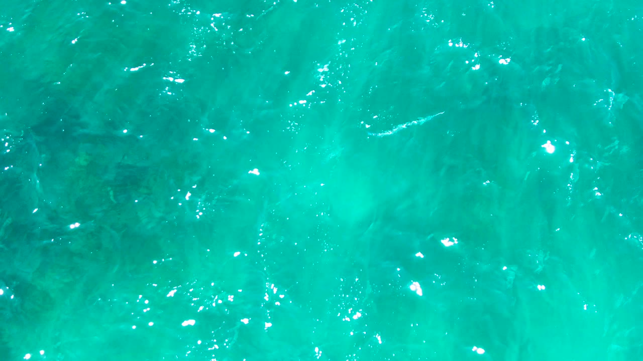Aerial turquoise water at Punta Buzzards shows a band of ripples, scattered white highlights, and darker underwater areas beneath the surface, no visible people.