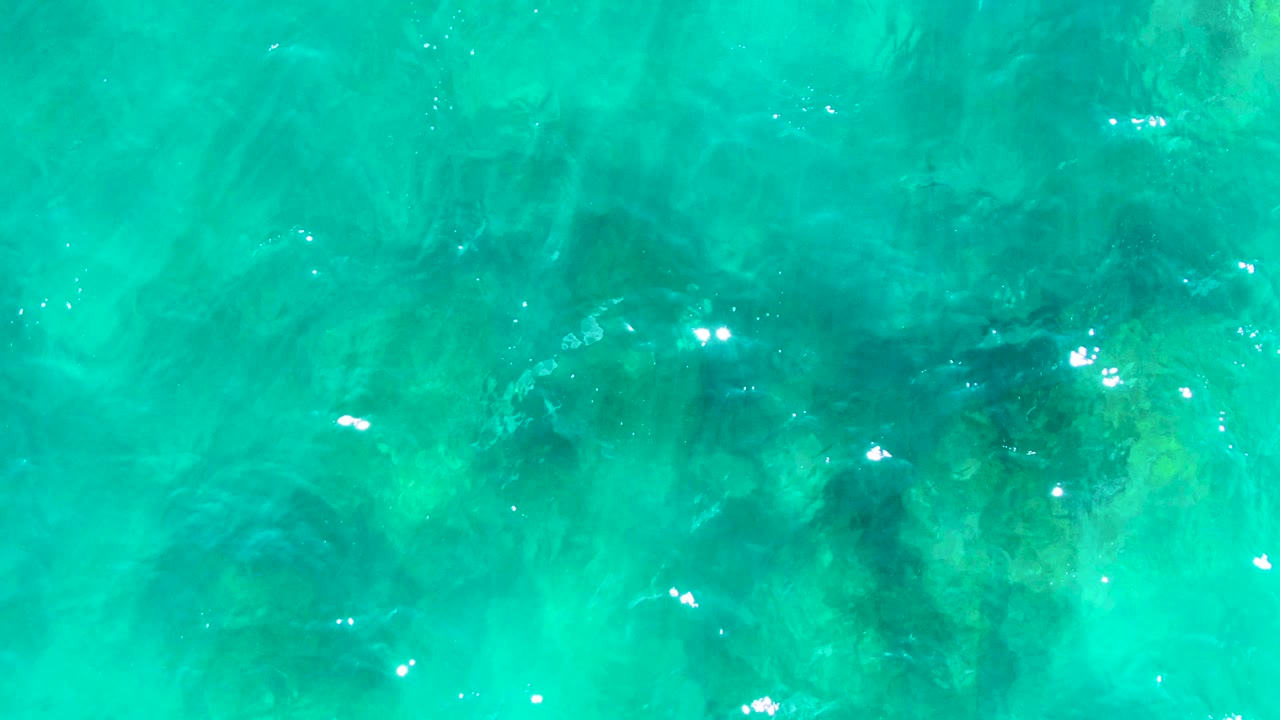 Top-down view of turquoise ocean water at Punta Buzzards, Baja California Sur, with dark underwater rocks and scattered bright reflections, no visible people.