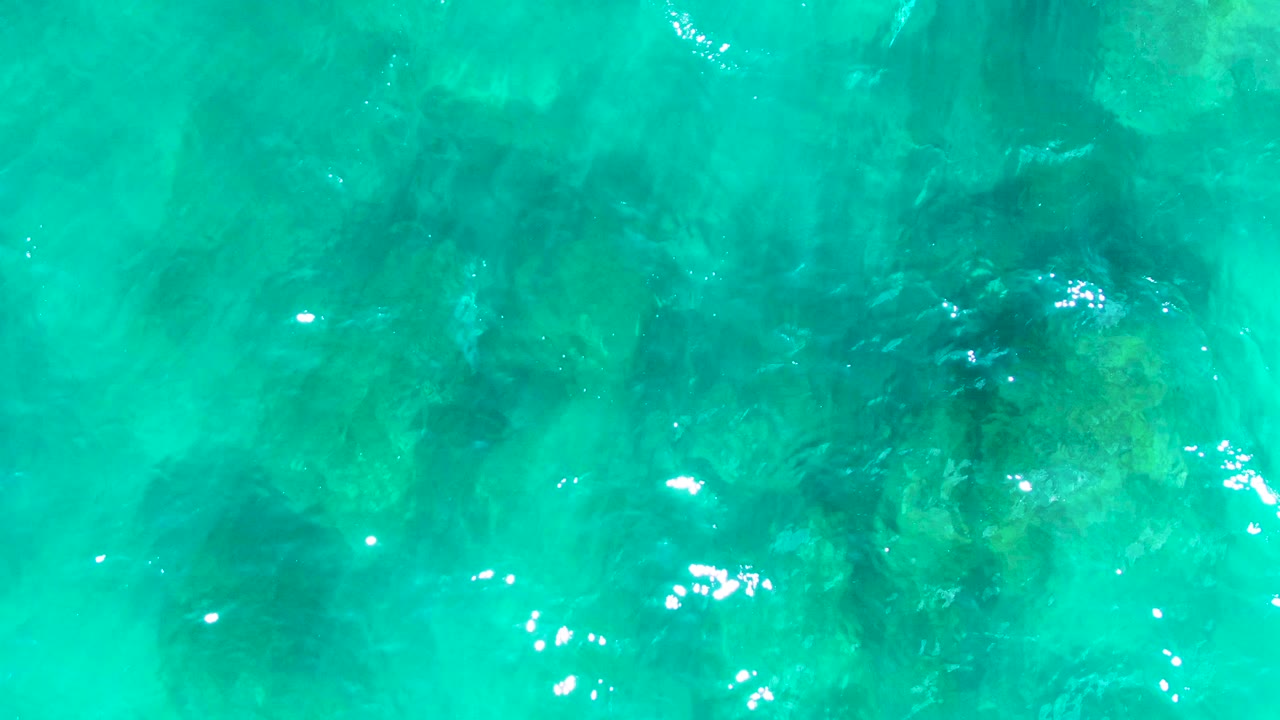 Clear blue-green ocean water at Punta Buzzards, Baja California Sur, with darker rocks visible below and small white light glints across the rippled surface, no visible people.