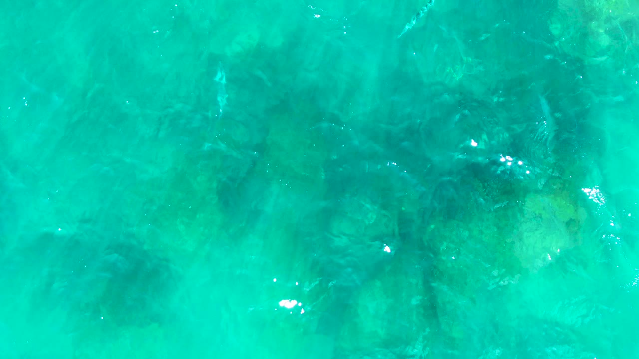 Turquoise ocean surface at Punta Buzzards, Baja California Sur, with dark reeflike underwater rock patches softened by ripples and no visible people.