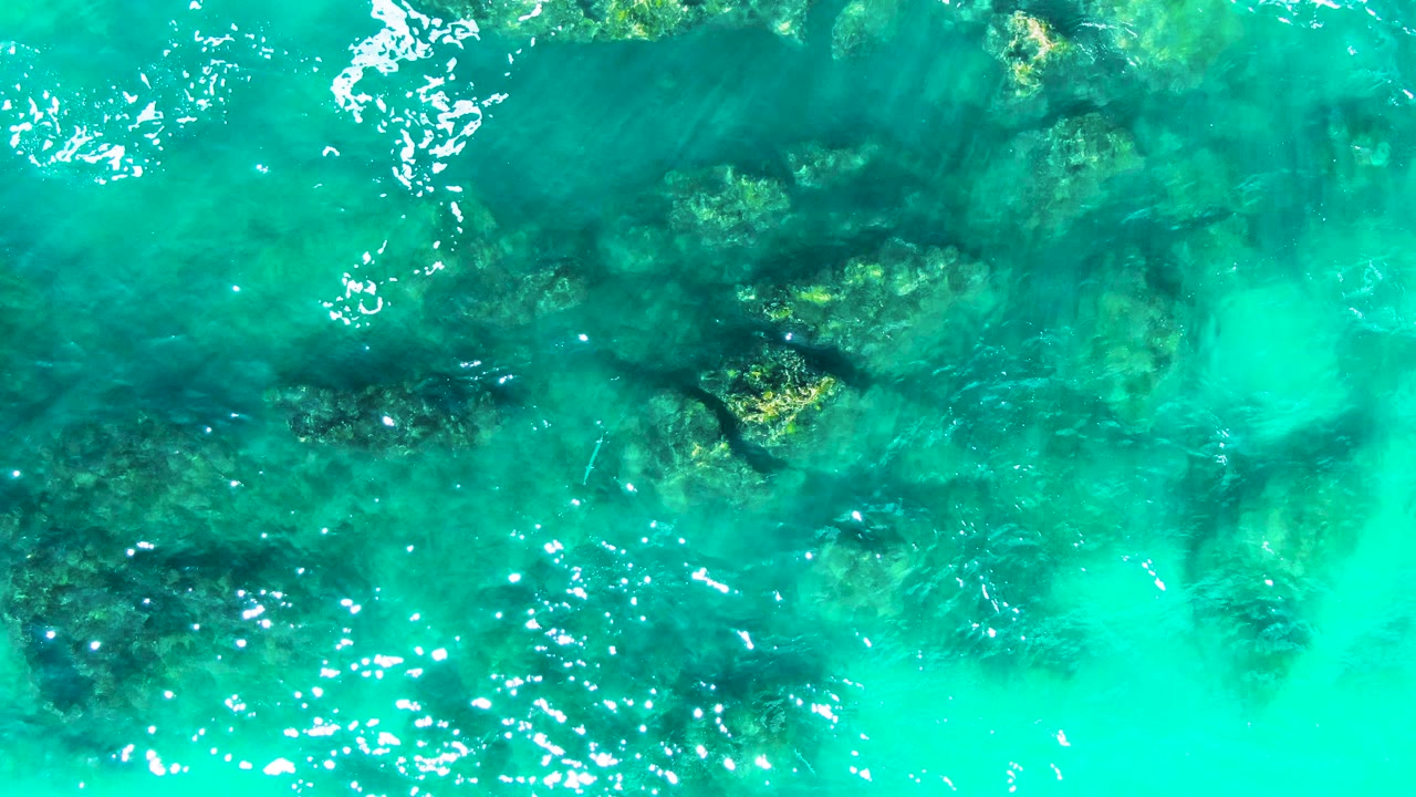 Shallow turquoise water reveals underwater rocks and foam trails.
