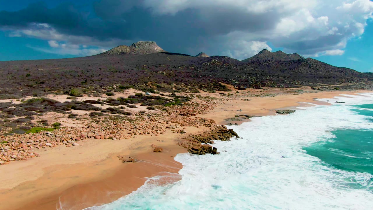 Opening aerial frame at Buzzards Beach in Baja California Sur, Mexico, showing white surf sweeping onto sand beside dark rocks, desert scrub inland, low hills, heavy clouds, and no visible people.