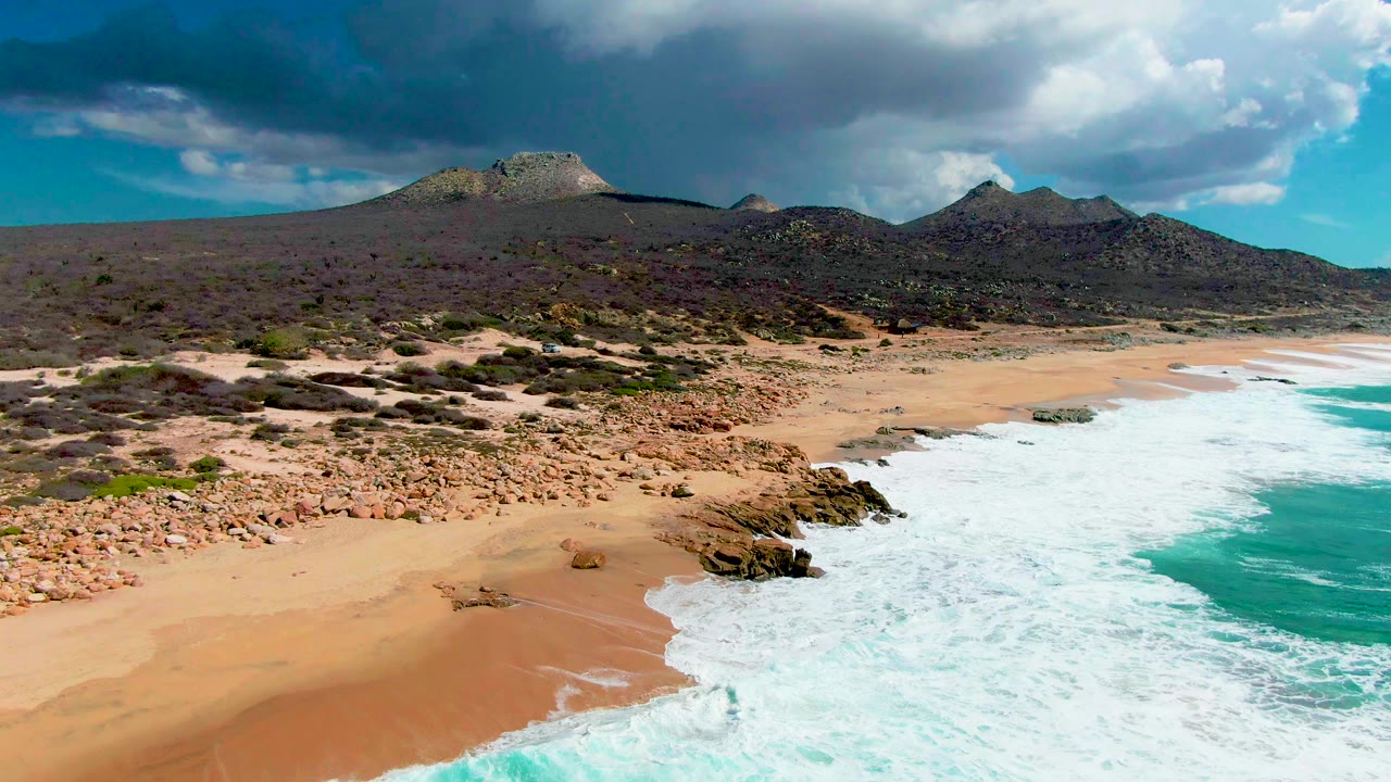 At 0:01, the aerial view shows foam widening across the right side of the sandy shore at Buzzards Beach, with rocky outcrops, scrubland, distant hills, large clouds, and no visible people.