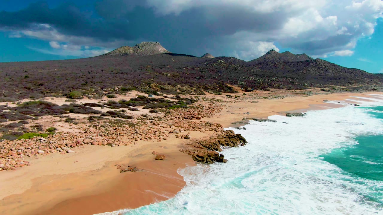 At 0:02, white water fills more of the nearshore edge while the sandy Buzzards Beach coastline curves past rocky points, desert vegetation, low hills, cloud shadow, and no visible people.
