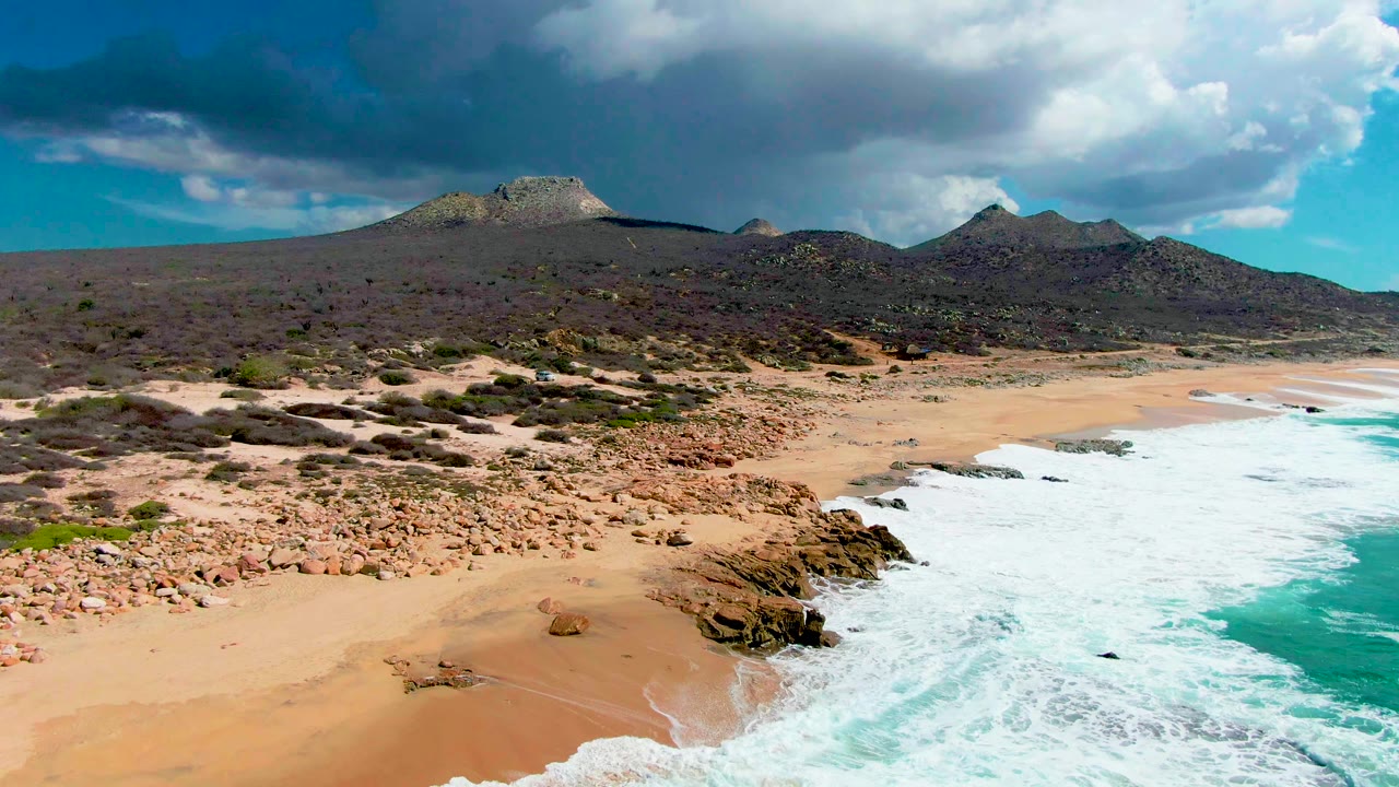 Surf recedes near rocky outcrops on an empty sandy beach below desert hills.