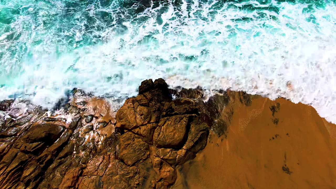 Overhead view at Buzzards Beach in Baja California Sur showing turquoise surf breaking into white foam over dark rocks and golden sand, with no visible people.