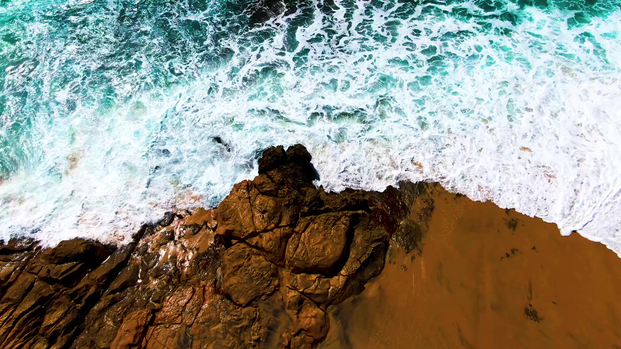 At Buzzards Beach in Baja California Sur, white surf spreads farther across the central rocks while a band of wet sand remains visible below, with no visible people.