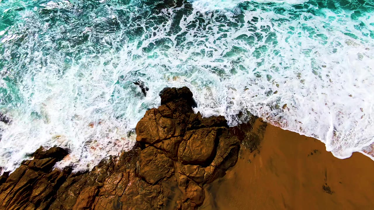 A top-down frame from Buzzards Beach in Baja California Sur shows a white breaking wave curling over the upper rocks and sandy edge, with no visible people.