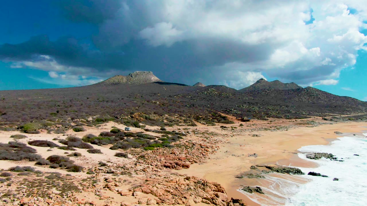 Wide rocky beach with receding surf, scrub, mountains, and clouds.