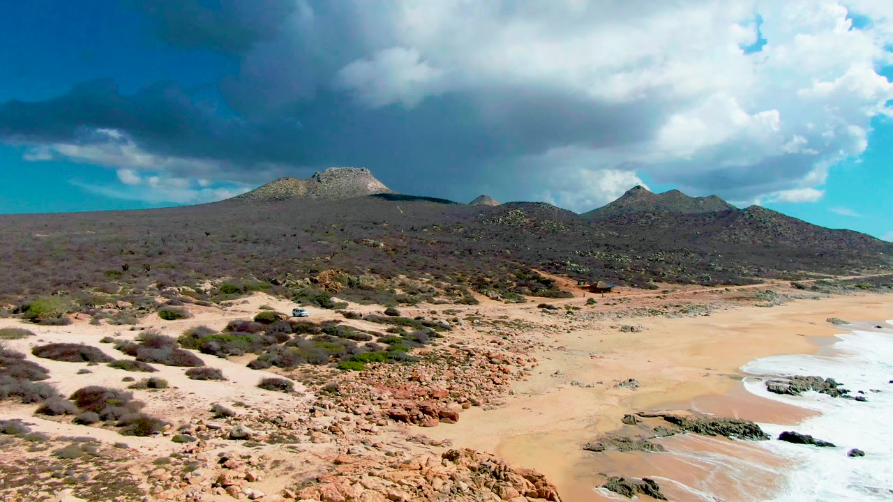 Buzzards Beach East Cape shoreline one second in, showing the foam line shifted along the right edge, pale sand in the foreground, rocky desert vegetation, distant mountains, and no visible people.