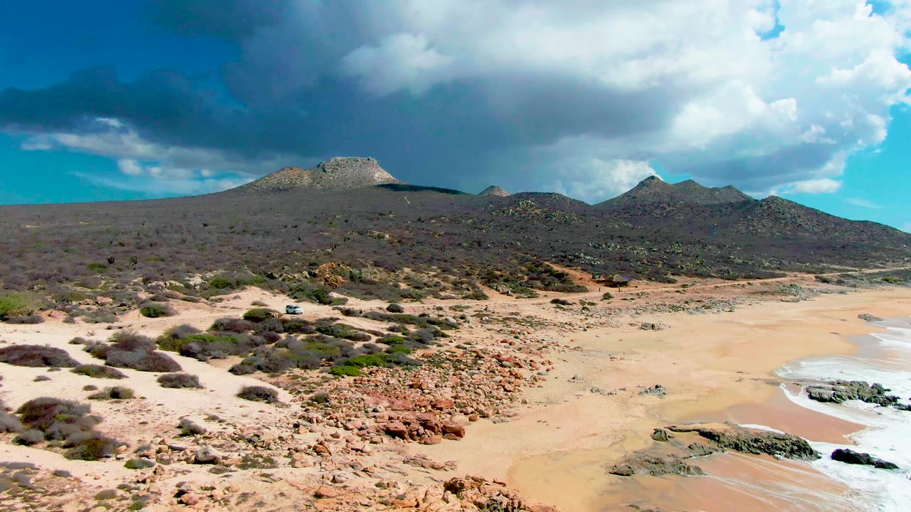Mid-clip view at Buzzards Beach East Cape with exposed sand, scattered rocks and scrub, distant brown mountain forms under large clouds, and no visible people.