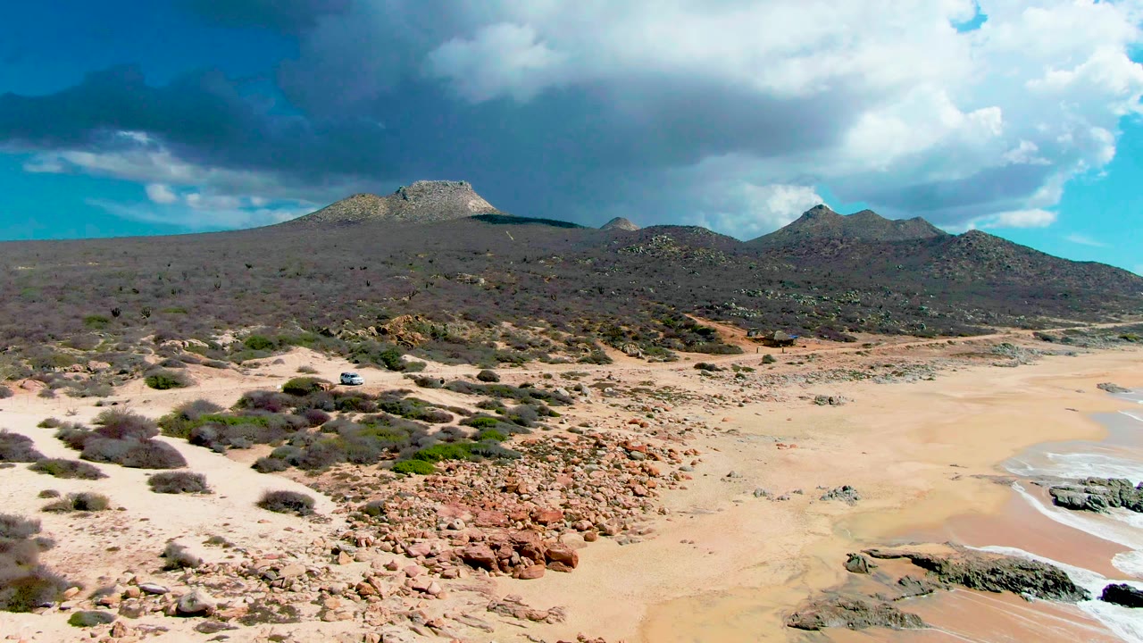 Later view of Buzzards Beach East Cape with more dry sand visible near the right shoreline, reddish rock patches in front, scrub across the slope, mountains in back, and no visible people.