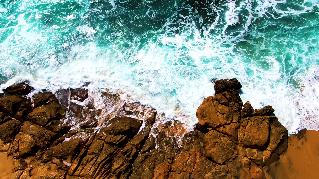 White surf washing over dark rocks and orange sand from overhead.