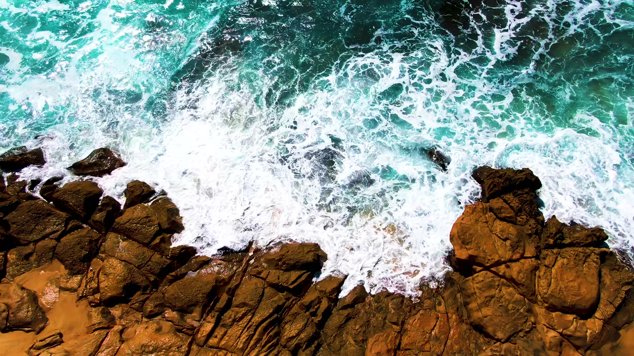 At 0:02, the overhead frame shows a bright band of breaking foam curling around larger brown rocks on the Punta Buzzards shore, with no visible people.