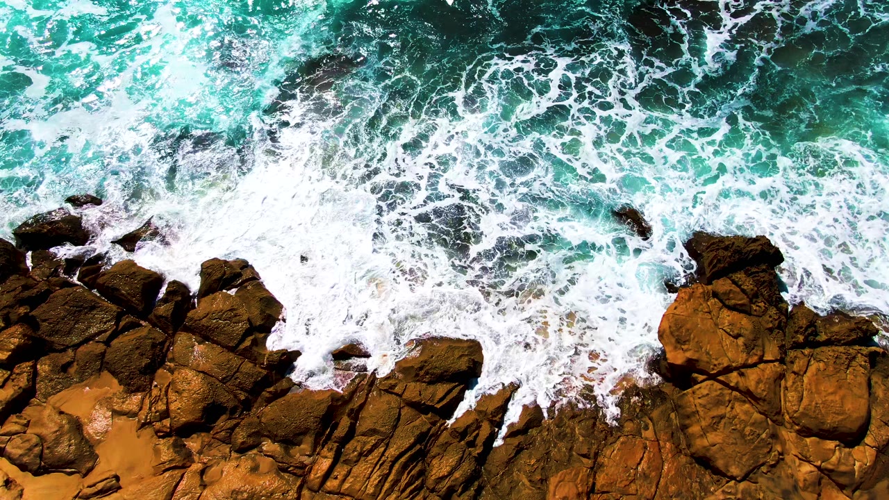 At 0:03, receding whitewater leaves more of the brown rock surface and orange sand visible along the Punta Buzzards shoreline, with no visible people.