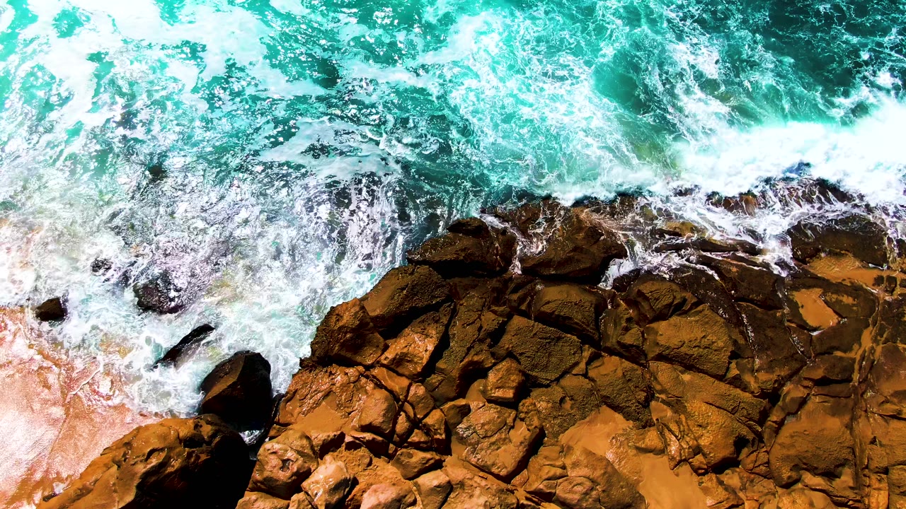 Turquoise waves breaking over brown coastal rocks and tan sand