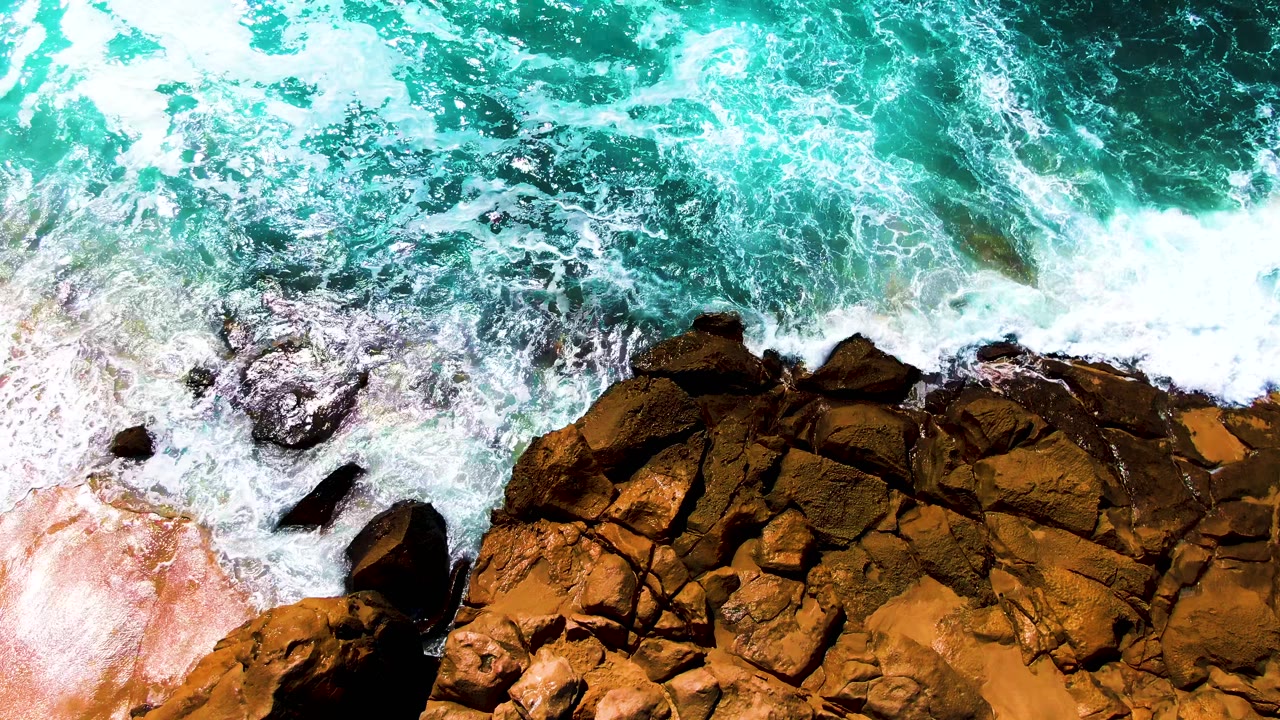 Aerial frame of white foam spreading across dark brown rock shelves under turquoise water at Punta Buzzards, with no visible people.