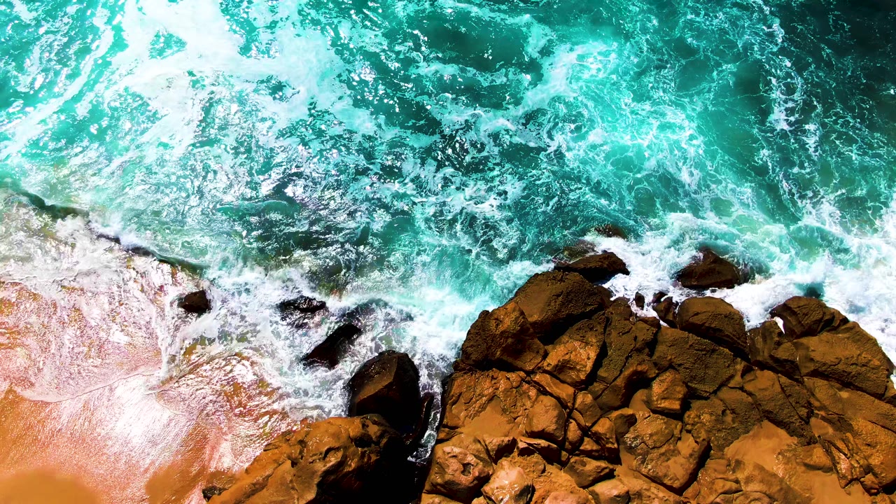 Aerial view of foamy wave wash surrounding brown rocks beside tan sand at Punta Buzzards, Baja California Sur, with no visible people.
