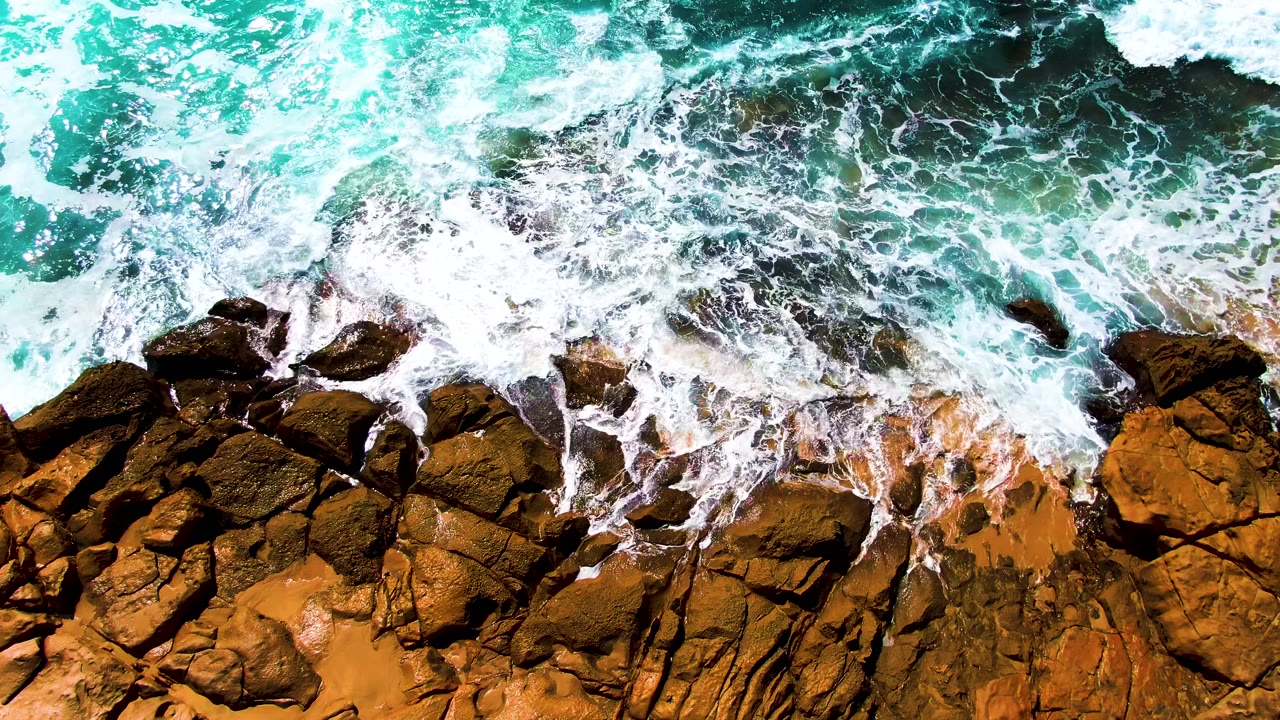 Overhead view of turquoise water and white foam reaching dark brown rocks at Punta Buzzards, Baja California Sur, with no visible people.