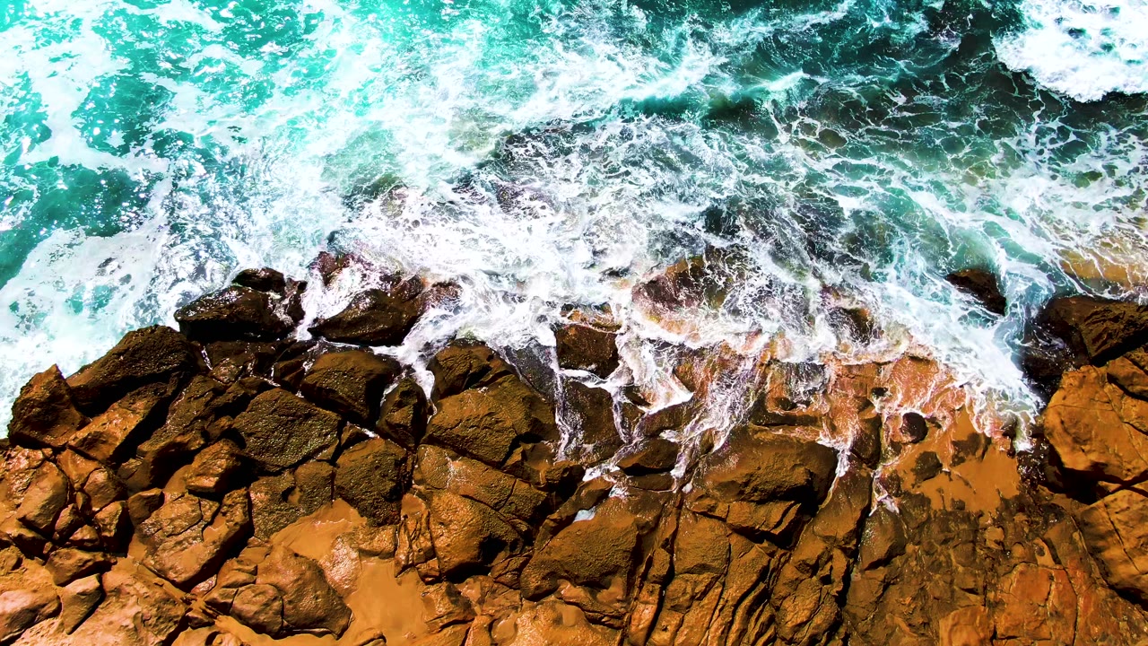 White foam spreads farther over jagged ochre and dark brown rocks as turquoise water churns above the Punta Buzzards shore, with no visible people.