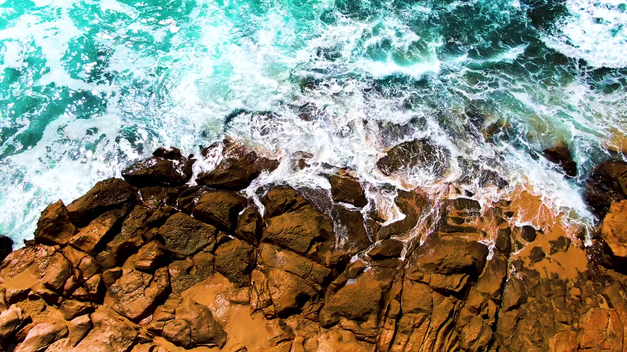The wave edge curls around several dark rock outcrops while ochre shoreline stone remains exposed below, with no visible people in the Punta Buzzards frame.