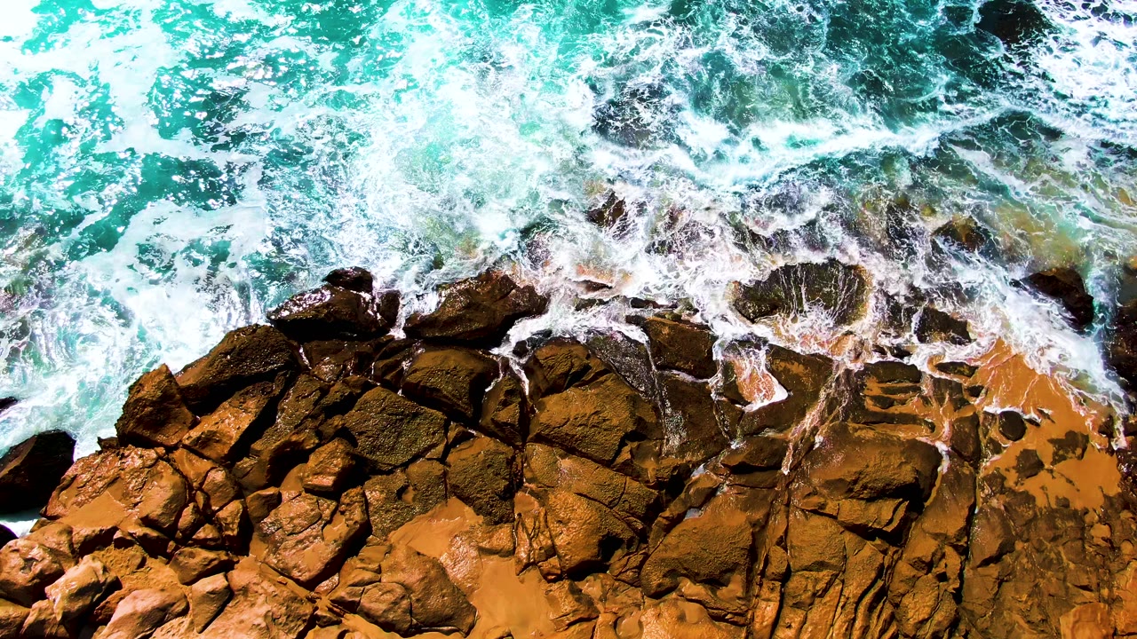 A broad band of white surf washes diagonally across rough brown rock, with turquoise water filling the upper half and no visible people at the shoreline.