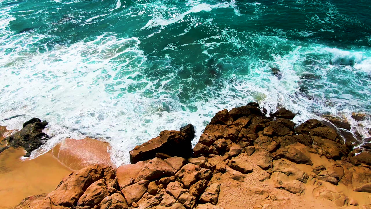 A wave pushes farther over the central rocks at Buzzards Beach, leaving white foam against turquoise water and orange sand, with no visible people.