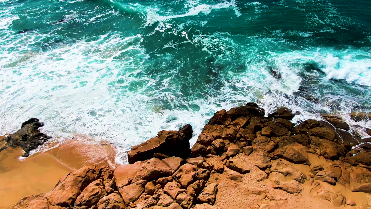 A broad line of breaking surf crosses the upper part of the frame above dark rocks and warm sand at Buzzards Beach, with no visible people.