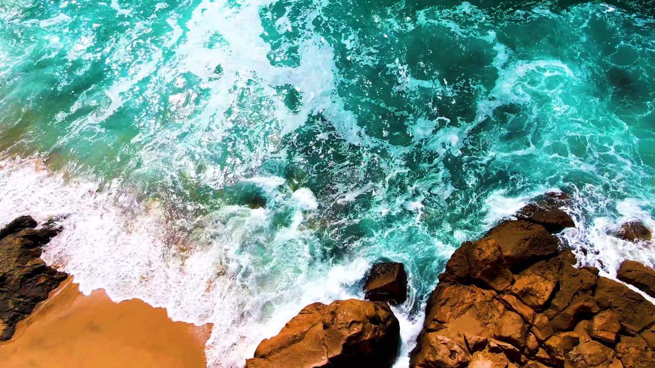 Overhead view at Buzzards Beach, East Cape, Baja California Sur, with turquoise water foaming over brown shoreline rocks and a strip of sand, no visible people.