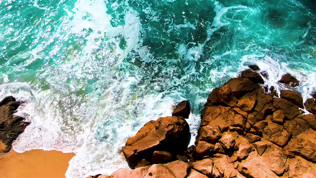Foam pushes into gaps between rust-brown rocks and a curved sandy edge, with blue-green water swirling above the shoreline in Punta Buzzards, no visible people.
