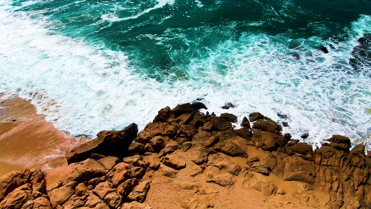 Aerial frame of white surf foam advancing over jagged brown shoreline rocks beside a small tan sand section at Buzzards Beach, with no visible people.