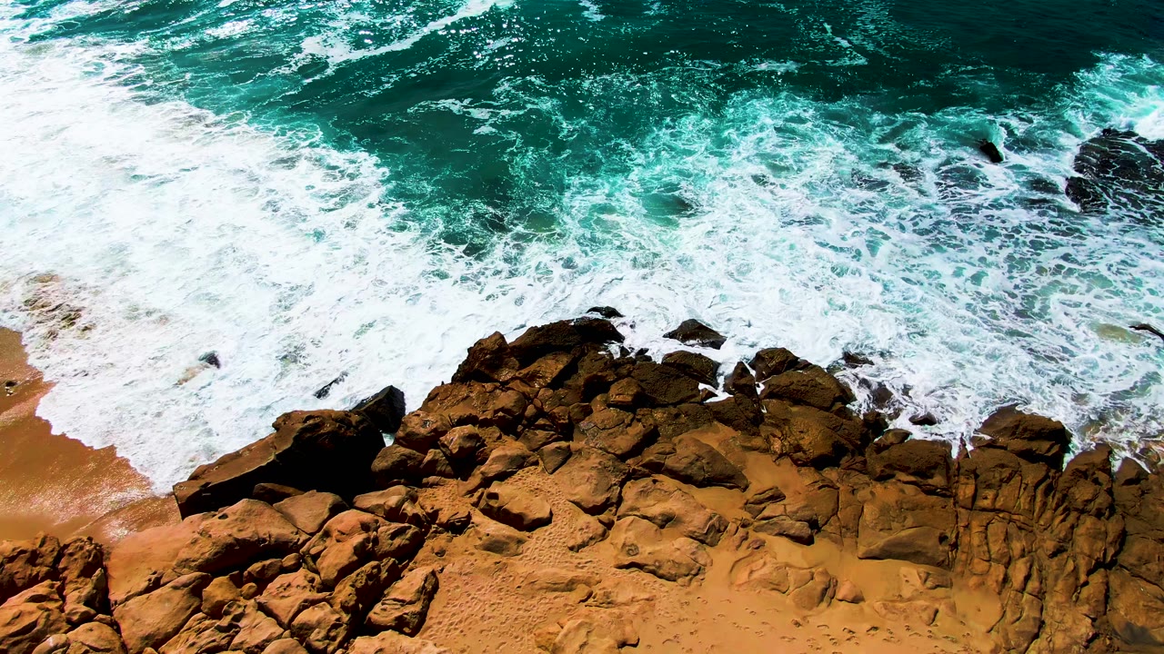 Overhead view of foamy turquoise water covering part of the rocky shore while tan sand remains visible along the lower left at Buzzards Beach, no visible people.
