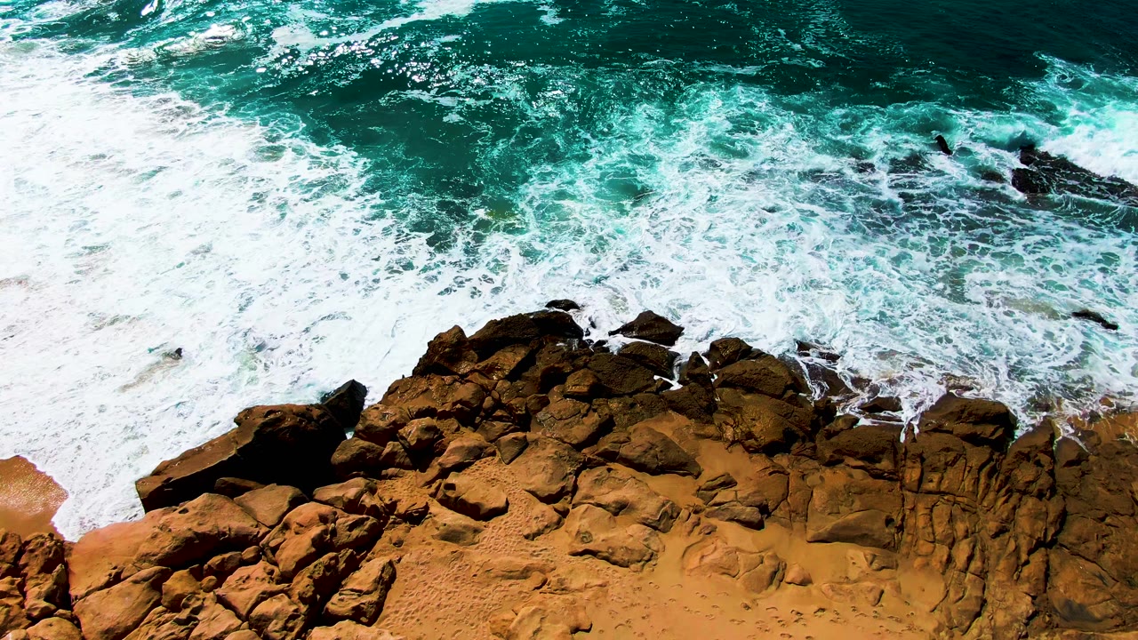 Aerial coastal view at Buzzards Beach with bright white foam sweeping diagonally over the rocks and sand while darker turquoise water sits offshore, no visible people.