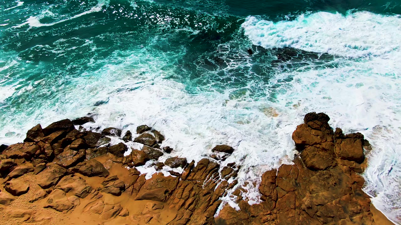 Overhead view at Punta Buzzards, Baja California Sur, where white surf breaks against orange-brown rocks below dark teal water, with no visible people.