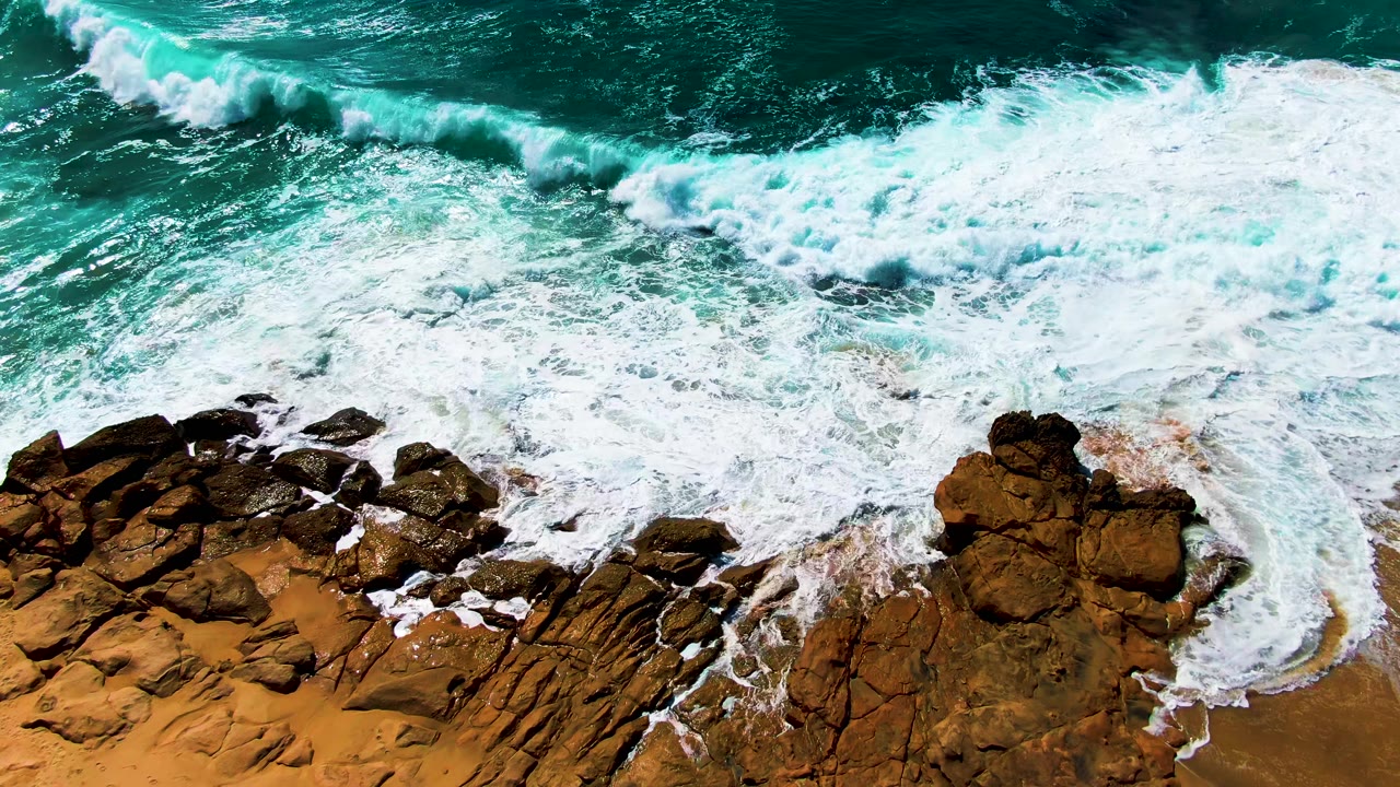 At Punta Buzzards, a broad band of whitewater spreads across the upper frame while jagged brown rocks and sand hold the lower edge, with no visible people.