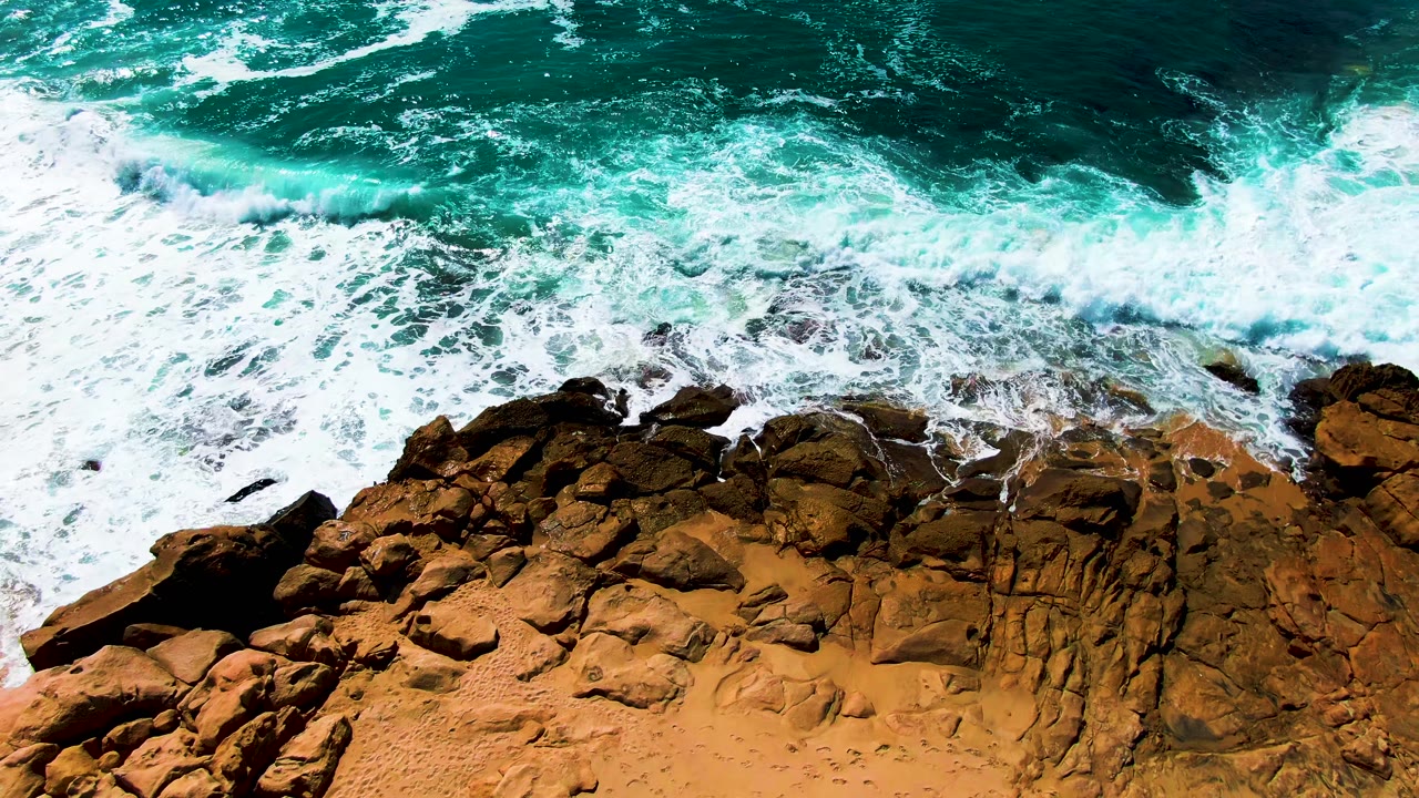 Top-down view of turquoise ocean and white foam meeting brown rocks at Punta Buzzards, Baja California Sur, with a sandy edge below and no visible people.