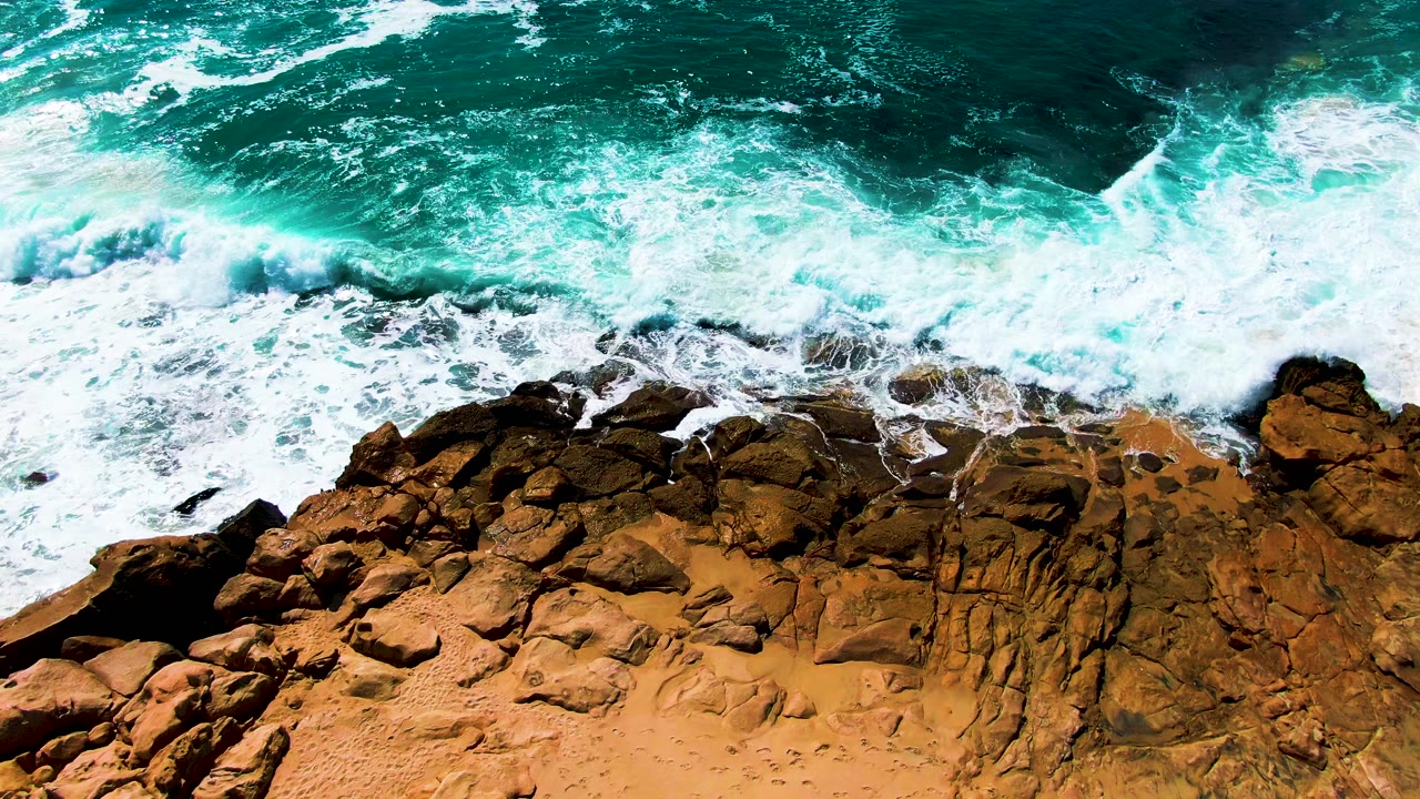 Aerial frame at Punta Buzzards showing a broad band of whitewater spreading over dark and ochre rocks, with turquoise sea above and no visible people.