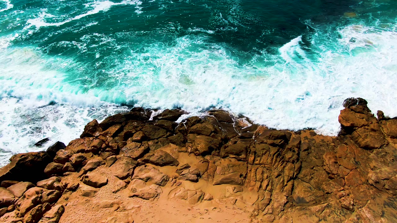 Overhead view of Punta Buzzards surf curling into white foam over jagged brown rocks, while deeper blue-green water fills the upper frame and no people are visible.