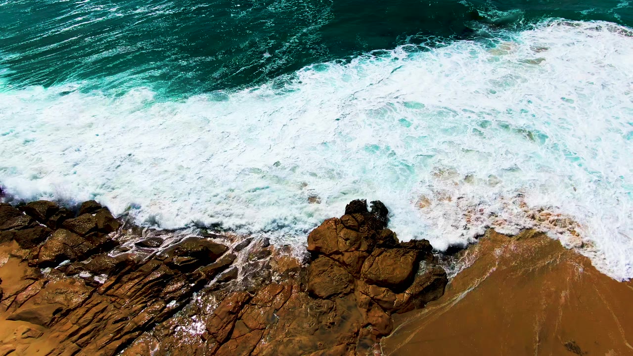 Overhead view at Buzzards Beach, Baja California Sur, with white surf pushing over dark shoreline rocks and wet sand, no visible people.