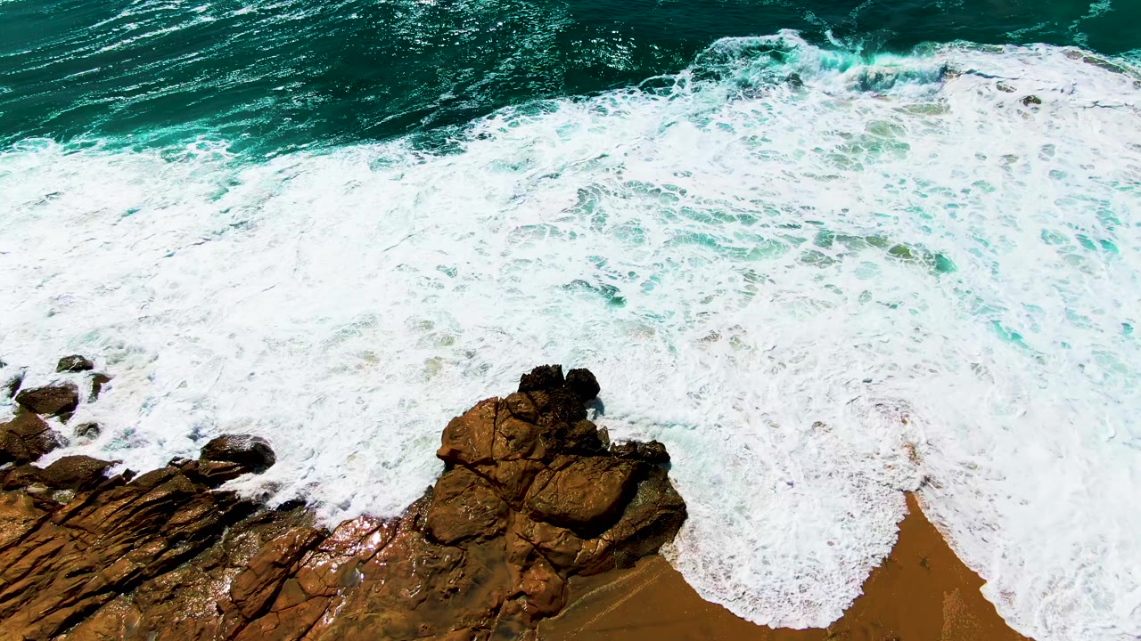 At Buzzards Beach in Baja California Sur, a broad sheet of white foam covers more of the rocky shoreline while turquoise water remains above, no visible people.