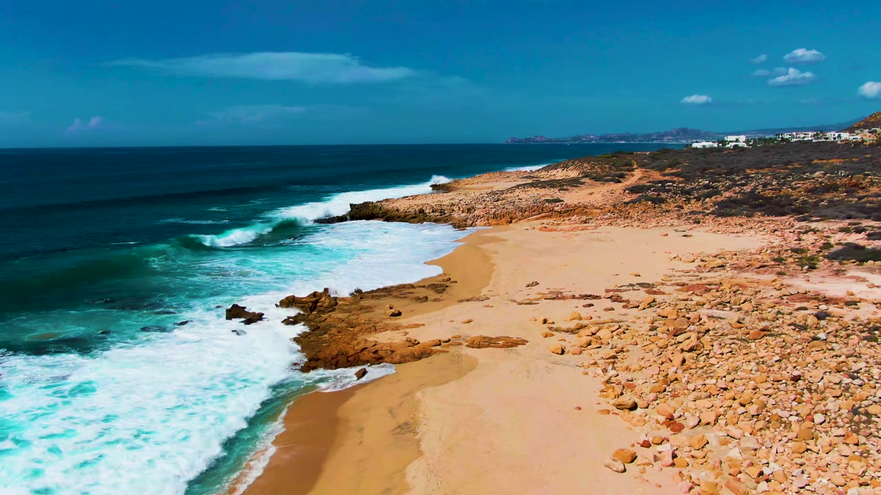 Aerial frame over Buzzards Beach East Cape BCS Mexico showing turquoise water, white foam, sand, rocky outcrops, sparse vegetation, distant buildings, blue sky, and no visible people.