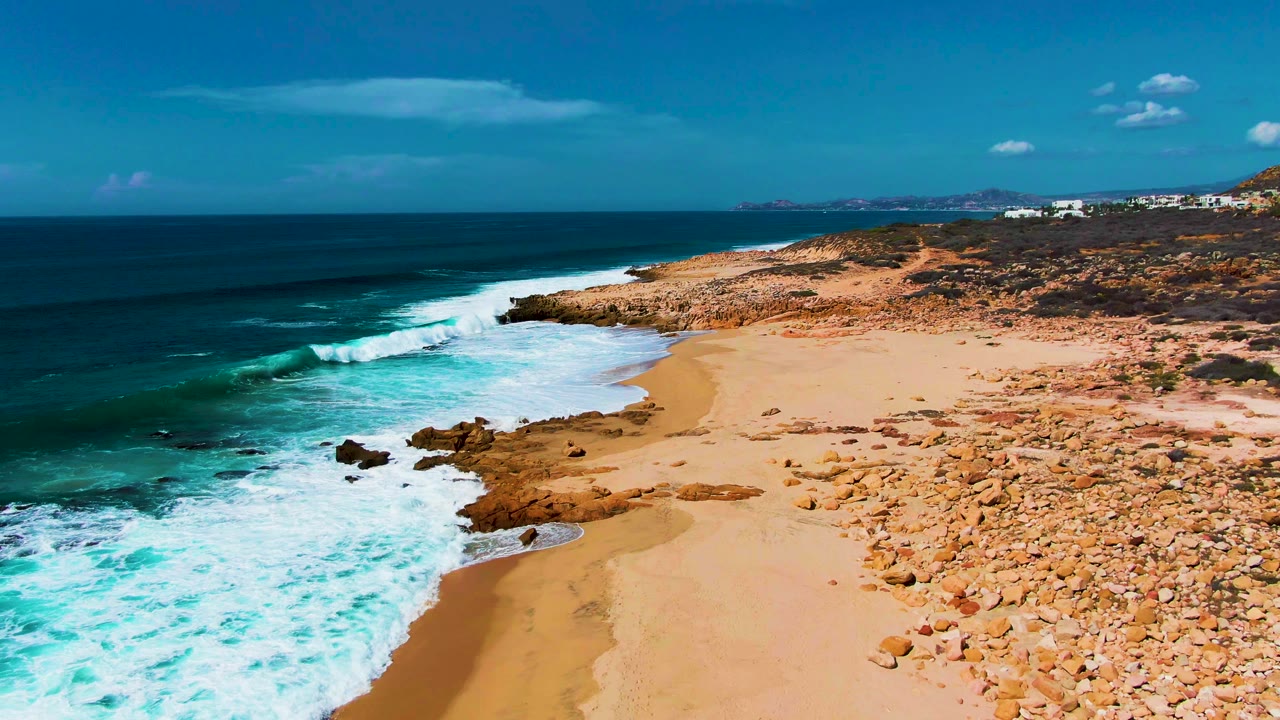 White waves break along a curving sandy shoreline with rocks inland.